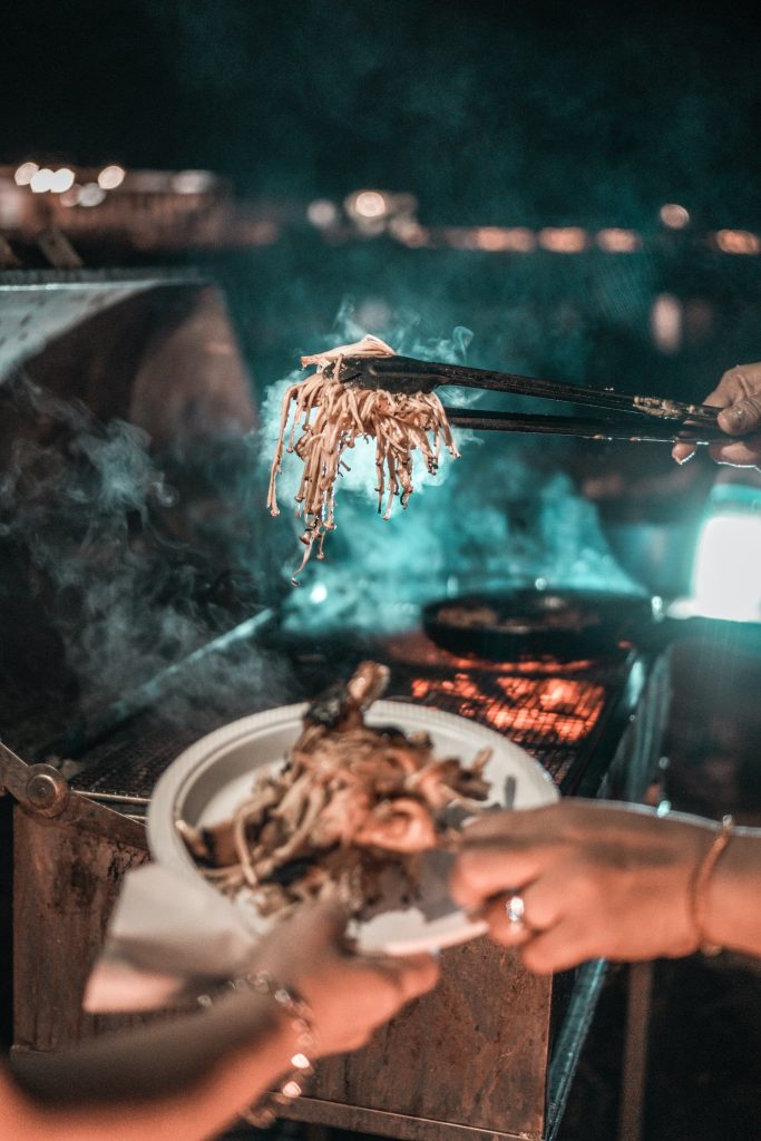 Balcony Gas Supply 1 Two people are grilling food on the balcony at night, with one person placing grilled noodles onto a paper plate held by another. The gas supply illuminates their faces in the smoky haze, and a bokeh background adds to the atmosphere.