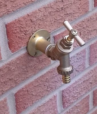 Balcony Tap 2 A brass balcony tap is mounted on a red brick wall. The faucet has a cross-shaped handle and is in the off position.