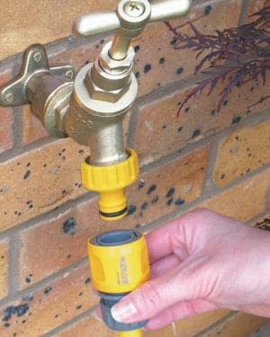 Balcony Tap 1 A hand holding a yellow and gray hose nozzle is about to connect it to a brass balcony tap installed on a brick wall.