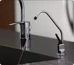 Under sink water filter 1 Close-up of two modern kitchen sink faucets, one standard with a lever handle and one taller with a curved spout, both mounted on a dark countertop. An under sink water filter system ensures that the water from these stylish faucets is always clean and safe to use.