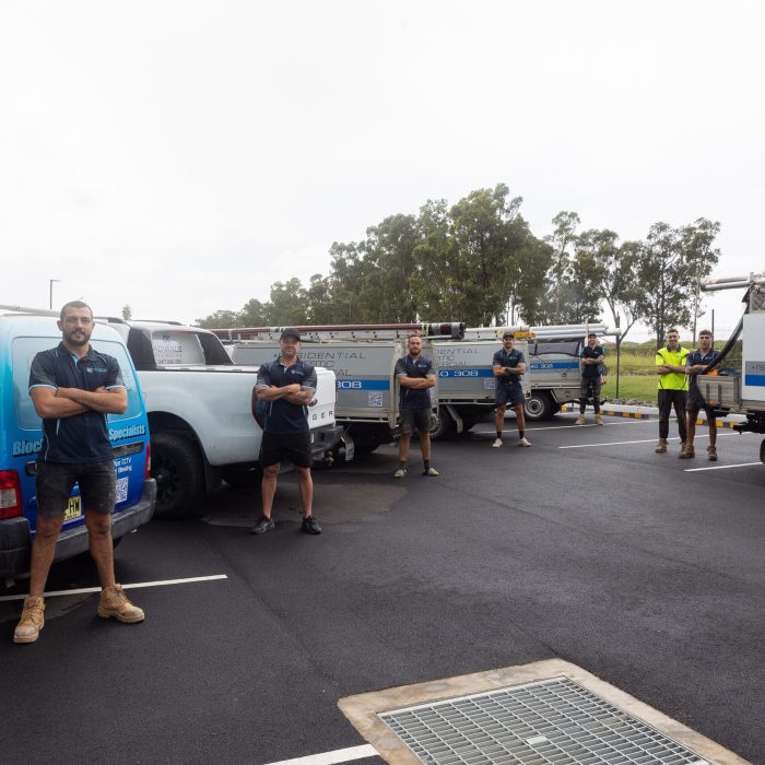A group of six people stand in front of three blue and white plumbing services trucks, parked in a lot. They are all wearing work clothing and appear to be posing for the photograph. A group of six people stand in front of three blue and white plumbing services trucks, parked in a lot. They are all wearing work clothing and appear to be posing for the photograph.